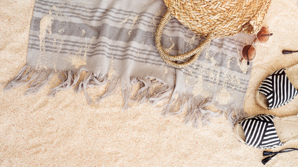 Flat lay of a beach towel, woven straw bag, sunglasses and striped sandals on sandy beach, representing summer essentials.