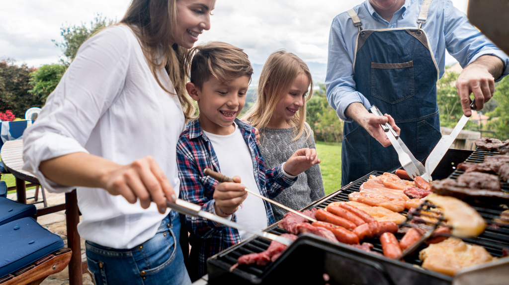 Smiling family enjoying a backyard BBQ, cooking sausages and chicken together on a grill during summer.